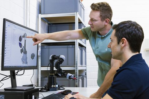 Zwei Wissenschaftler arbeiten am Rechner. Im Hintergrund steht ein Roboter