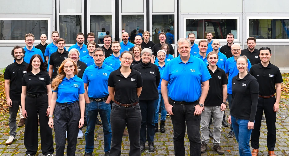 Eine größere Gruppe von Mitarbeitenden steht in mehreren Reihen im Freien vor einem modernen Gebäude mit großen Fenstern. Etwa die Hälfte trägt leuchtend blaue Polo-Shirts, die andere Hälfte schwarze. Alle blicken zur Kamera; einige lächeln. Der Boden ist mit herbstlichem Laub bedeckt, was einen farbigen Kontrast zu den einheitlichen Shirts bildet.