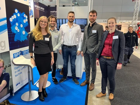 The image shows a group of five people at a trade fair or conference, posing in front of an information booth. The booth has a blue carpet, and several posters and displays are visible in the background. One of the posters features a map of Germany with various markers, along with the text “SPP 2013” and “Academic research on the metal industry” The individuals are wearing name tags and are dressed formally.