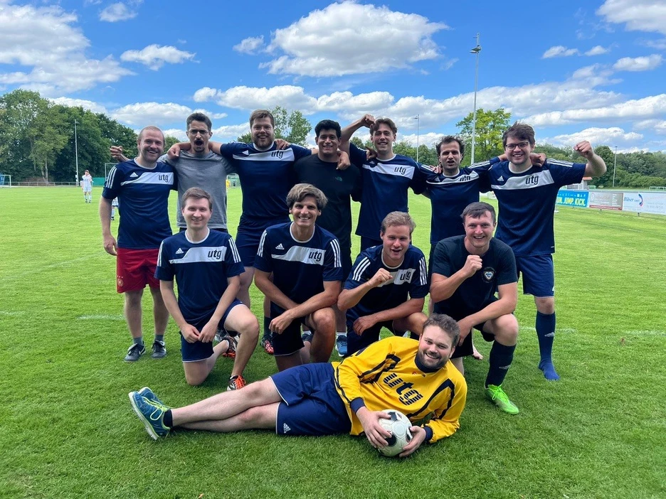 Das Bild zeigt 12 junge Männer in dunkelbleuen Fußballtrikot mit dem weiß hinterlegten Logo des utg. Die Personen schauen alle in die Kamera. Sie stehen als Mannschaft in drei reihen gruppiert auf einem Fußballrasen.