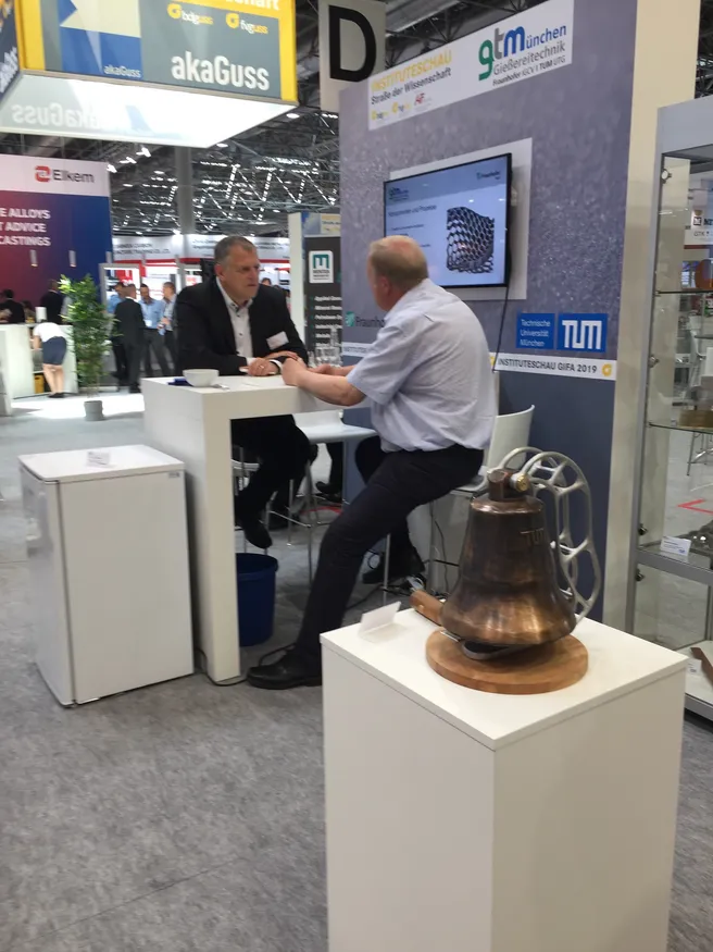 Prof. Volk im Gespräch mit einem Messebesucher am Stand der Gießreitechnik München. Im Vordergrund ist die Jazz-Glocke zu sehen, welche im Rahmen einer Masterarbeit am Lehrstuhl gegossen wurde.