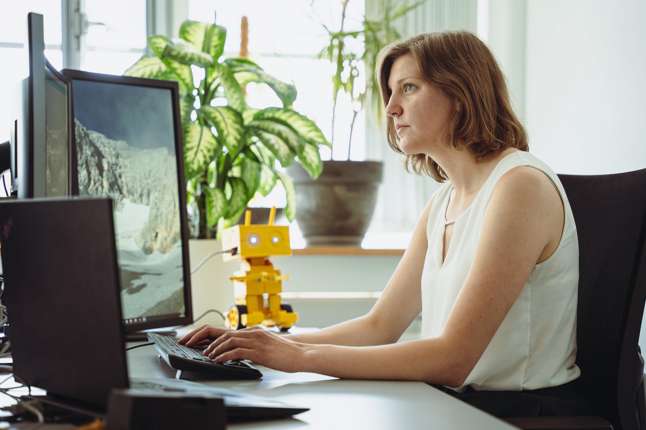 A woman is working at two screens. In the background is a robot
